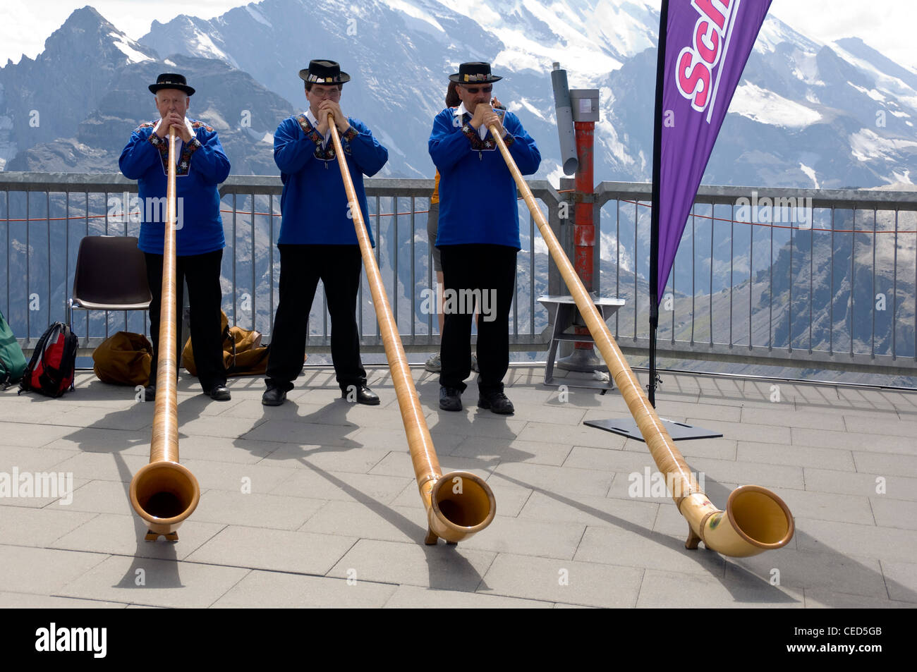 Horizontal close up portrait of three traditionally dressed Swiss men playing the Alpine horn with the Swiss Alps behind them. Stock Photo