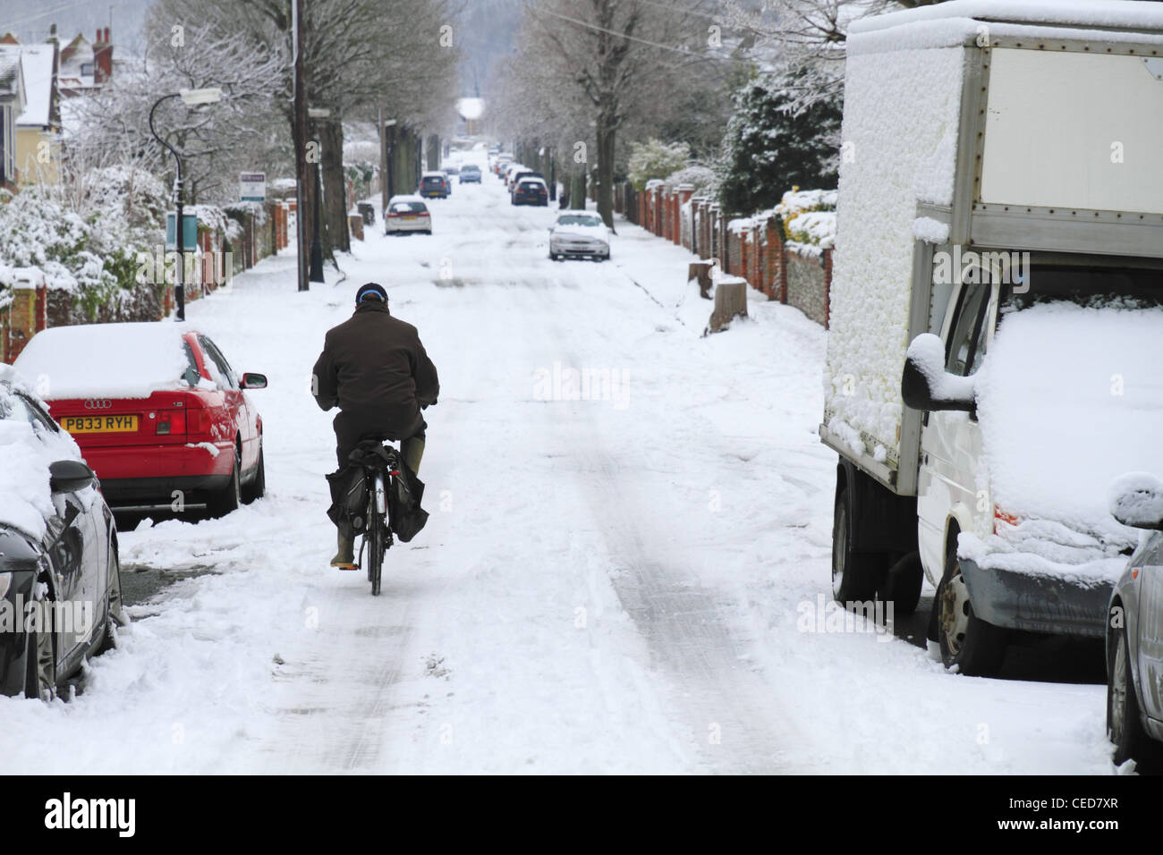 An old elderly man person cycling his bike bicycle uphill up hill in the snow on a snowy street road Eastbourne UK Stock Photo