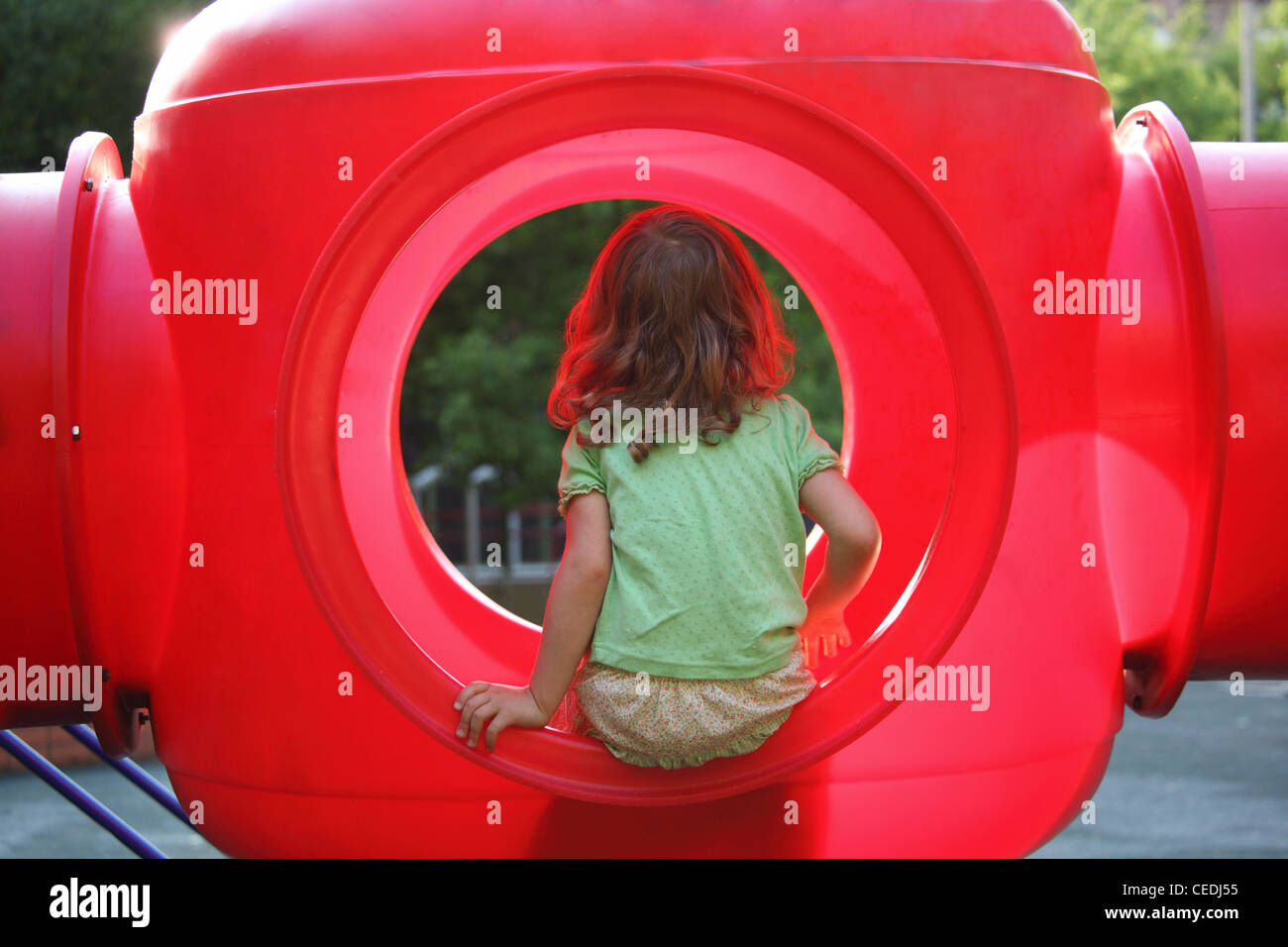 little girl sits in red plastic box on playground, view from back Stock Photo
