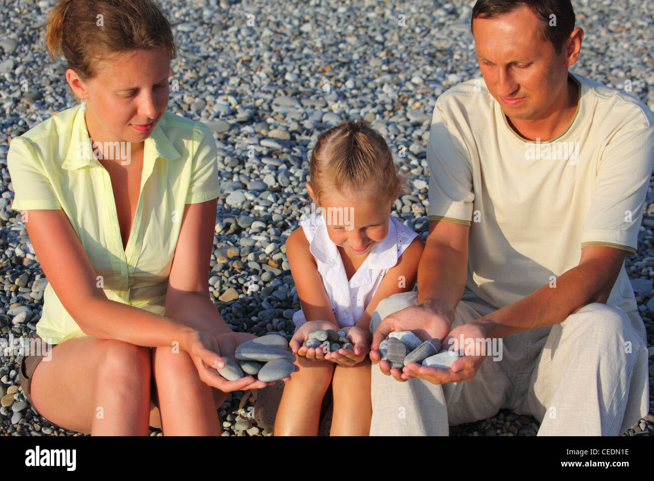 Parents and daughter sits in pebbly beach ind holding pebbles in hands Stock Photo