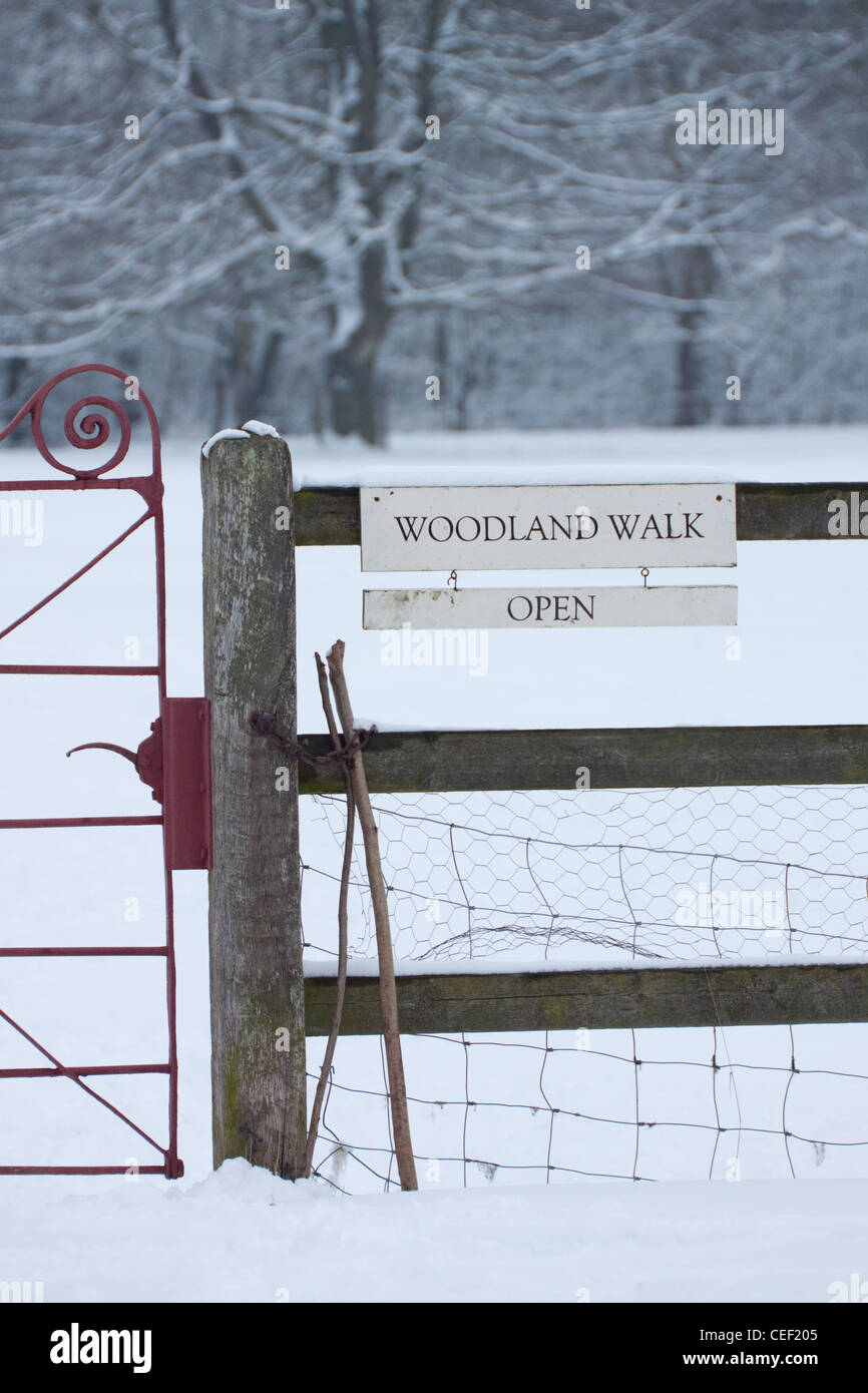A red metal gate into a snowy meadow in winter, with a sign on a wooden fence stating Woodland Walk Open Stock Photo