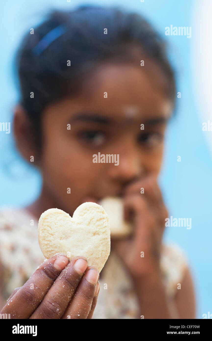 Young poor lower caste Indian street girl holding and eating an indian sweet heart shaped biscuit. Selective focus. Stock Photo