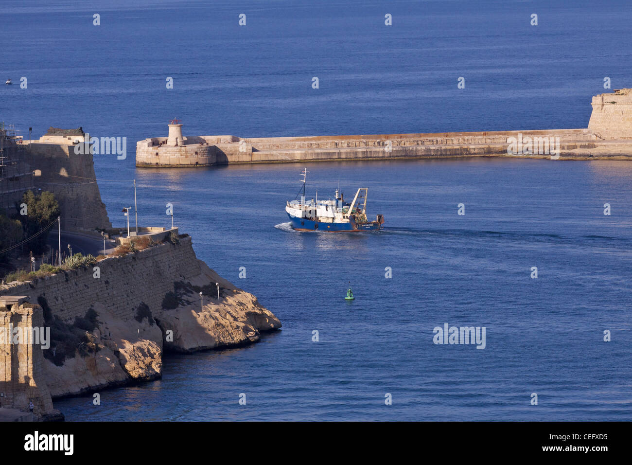 The lighthouse at the end of Ricasoli Point at the entrance to the Grand Harbour with a trawler heading out to sea at sunset Stock Photo