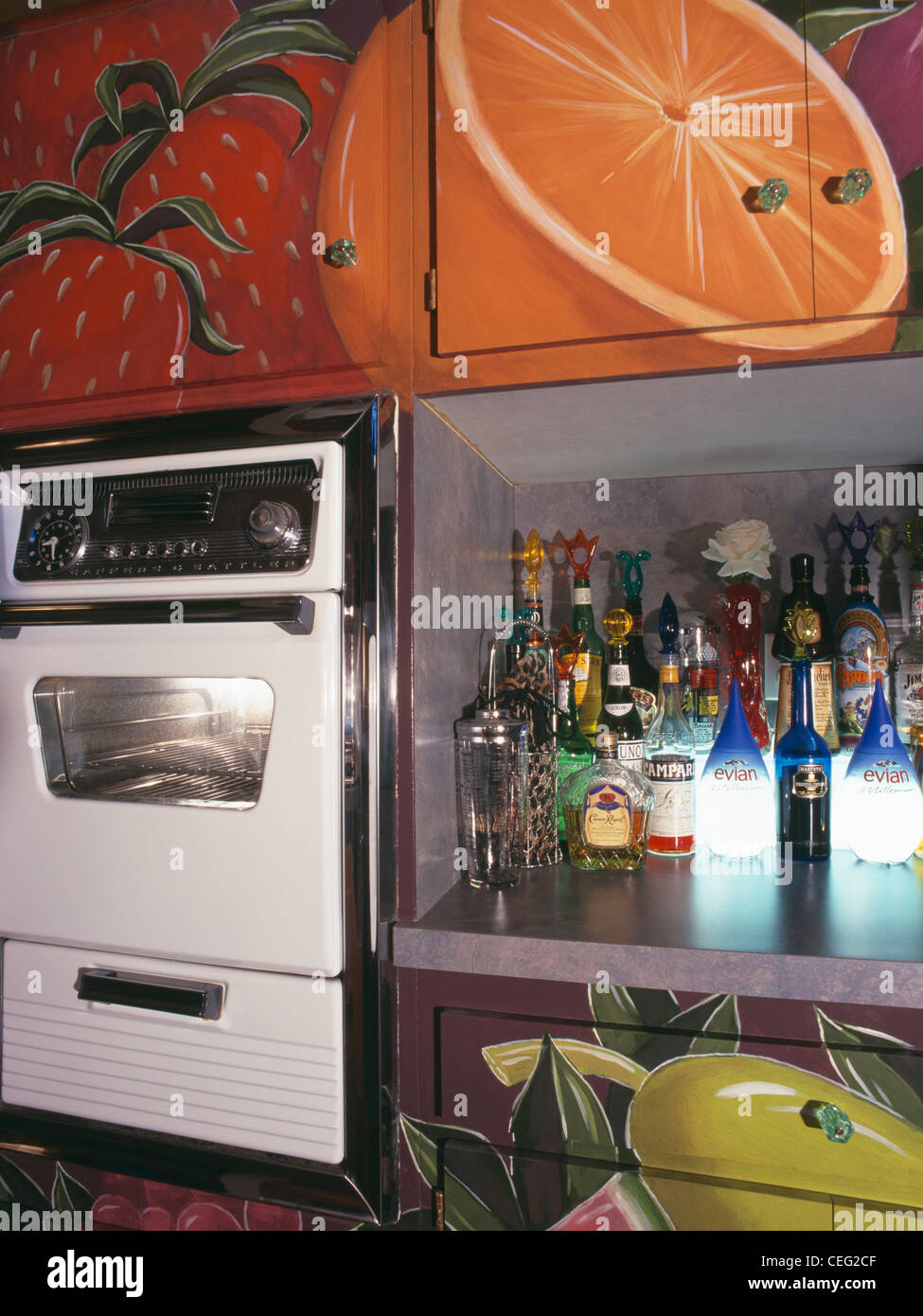 Close-up of oven and chilled drinks cabinet below trompe-l'oeil fruit on fitted cupboards in retro kitchen Stock Photo