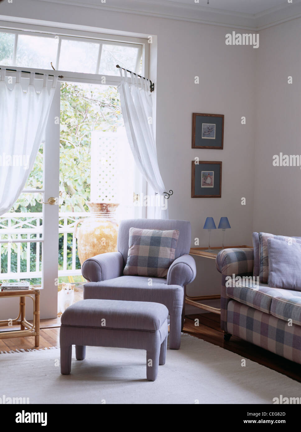 Blue armchair and footstool in front of French windows with white voile curtains on portiere rods Stock Photo