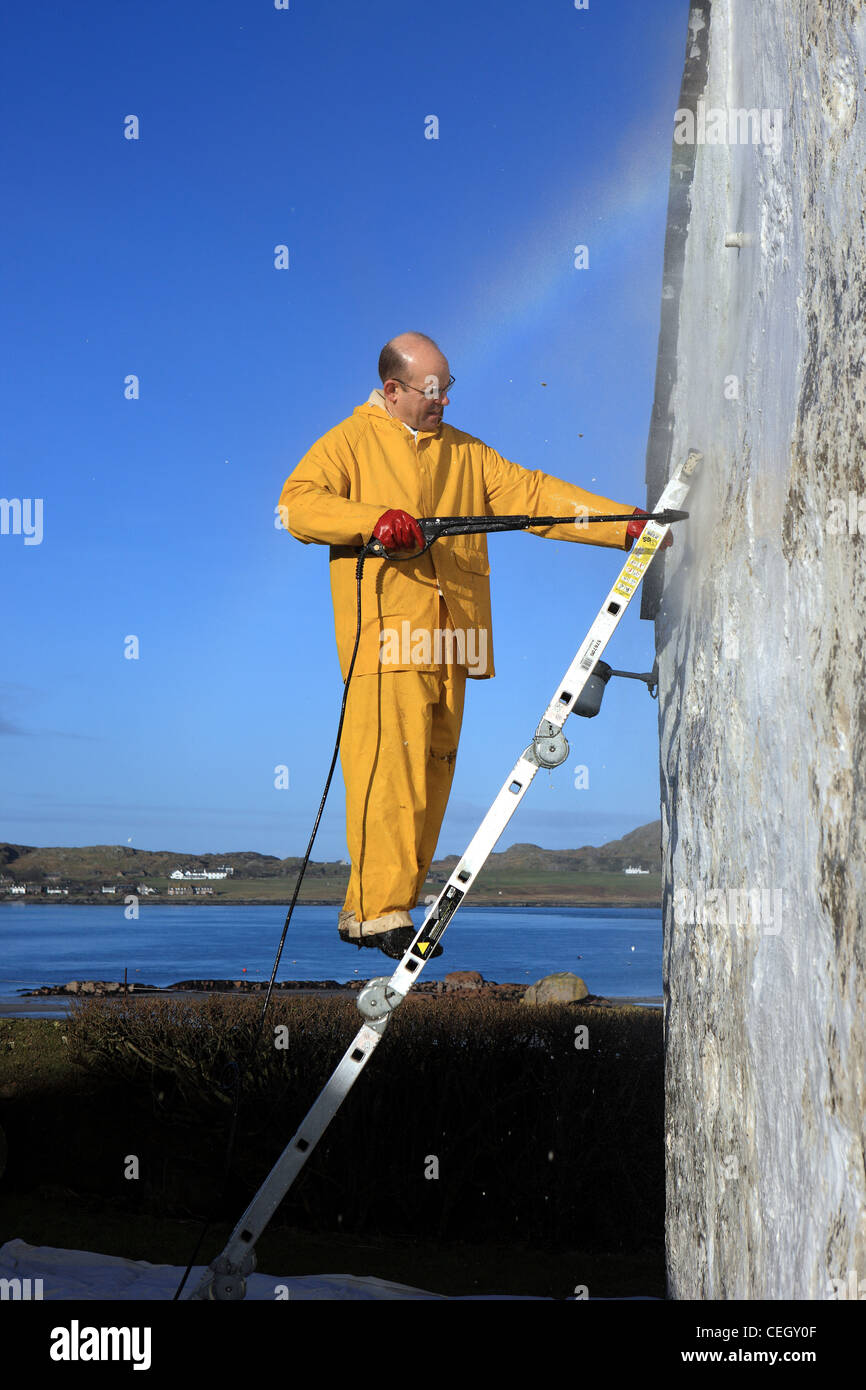 Man up a ladder using pressure washer to remove paint from a white