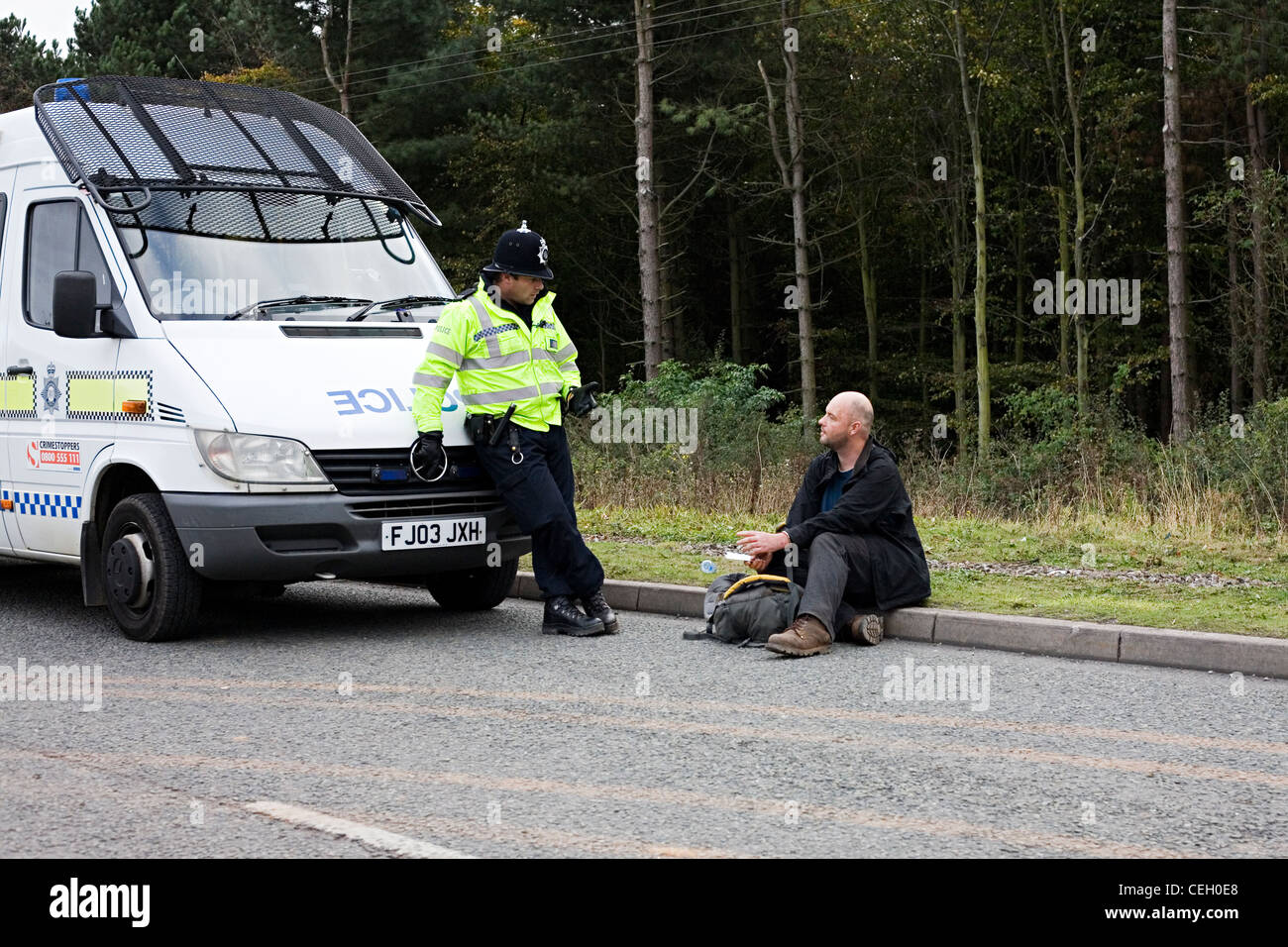 Man under arrest sat on the side of the road next to a police officer and a riot van. Stock Photo