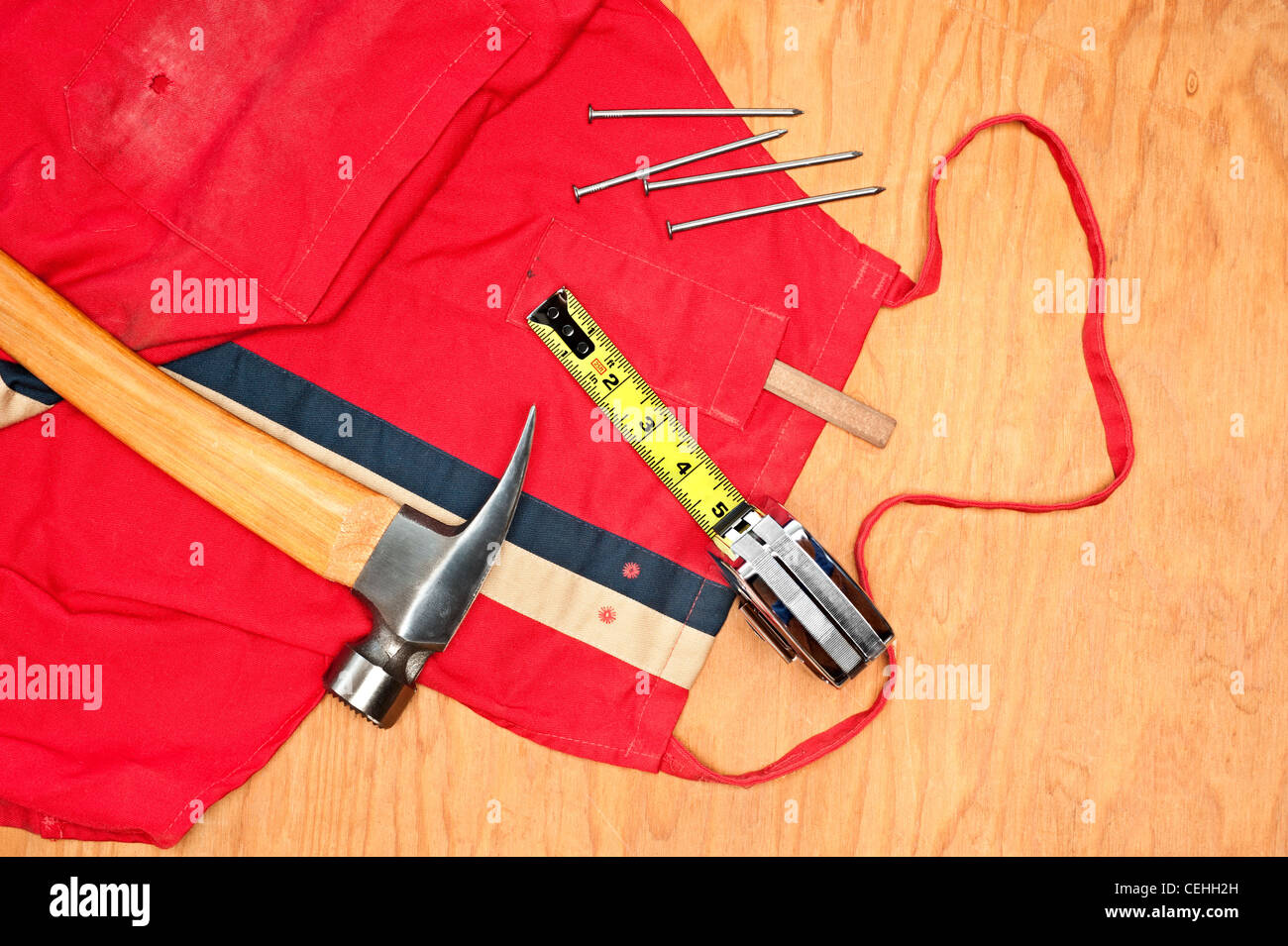 Woodworking tools including a hammer, tape measure, carpenters pencil and old red apron on a piece of plywood plank Stock Photo