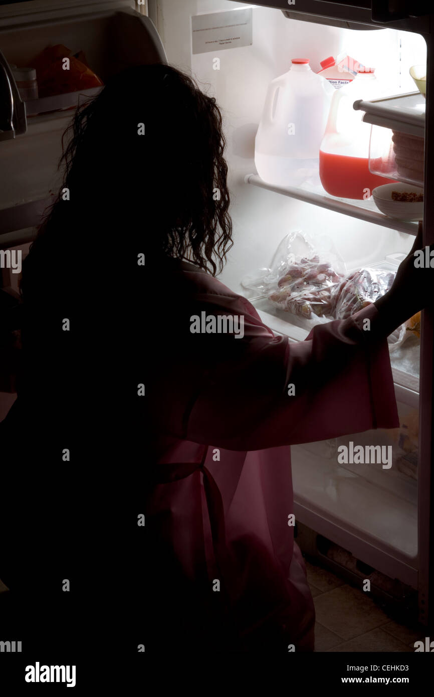 silhouette of overweight ethnic African American woman in pajama squatting by refrigerator looking for food in the night. Stock Photo