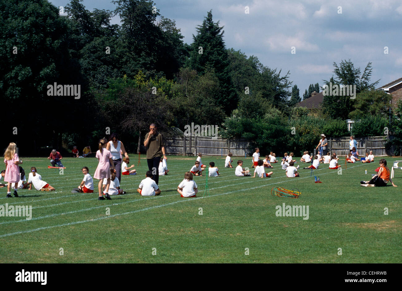 Primary School Sports Day Children Sitting On Grass With Teacher Holding A Whistle Stock Photo