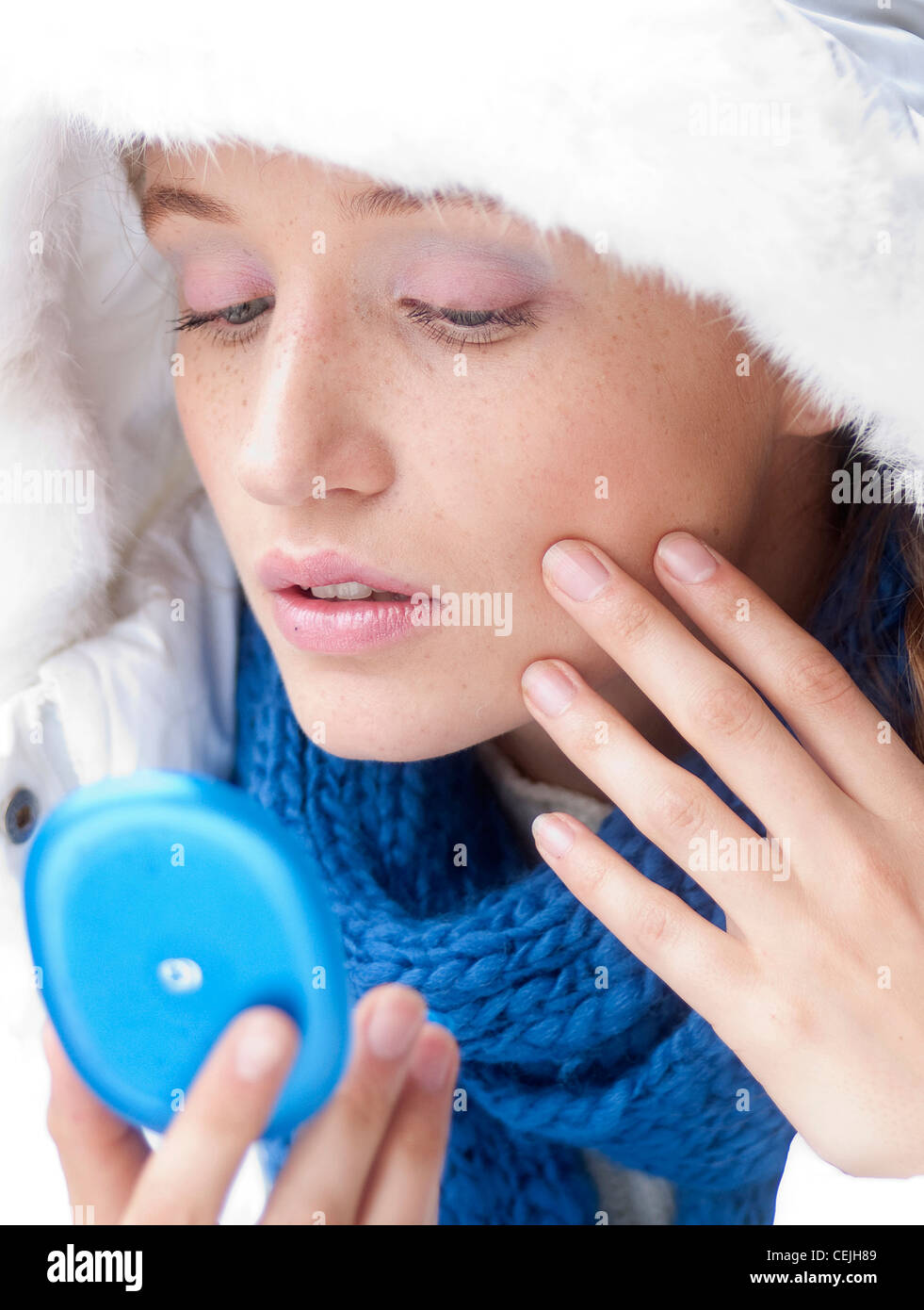 Female fair hair off her face, wearing a pink eyeshadow, white hooded parka and blue scarf, unsmiling, looking into a blue Stock Photo