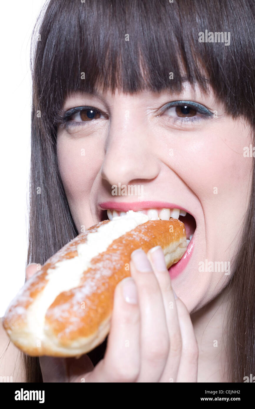 Female long fringed brunette hair, wearing green metallic eyeshadow and pink lipgloss, eating eclair, smiling, looking at Stock Photo