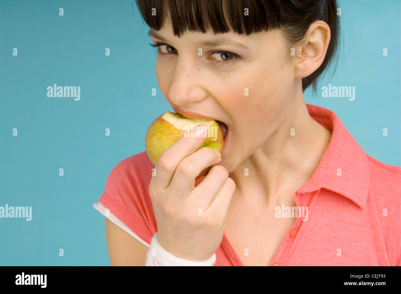 Female wearing red t shirt with open collar and white wristband taking bite from apple mouth open looking to camera  RBO Stock Photo