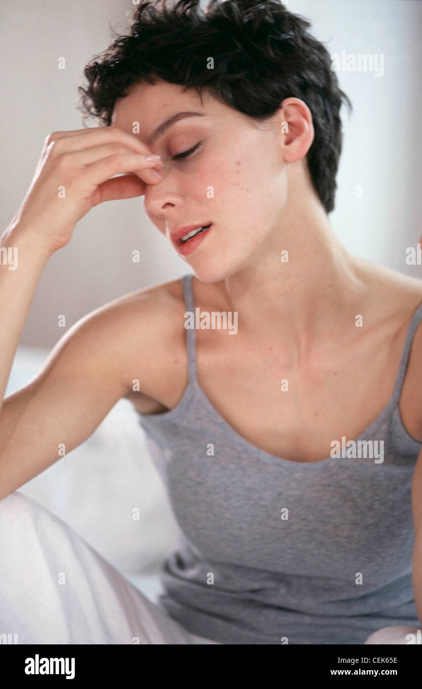 Semi profile of female short brunette hair wearing grey vest top and cropped white trousers, sitting on bed white sheet Stock Photo