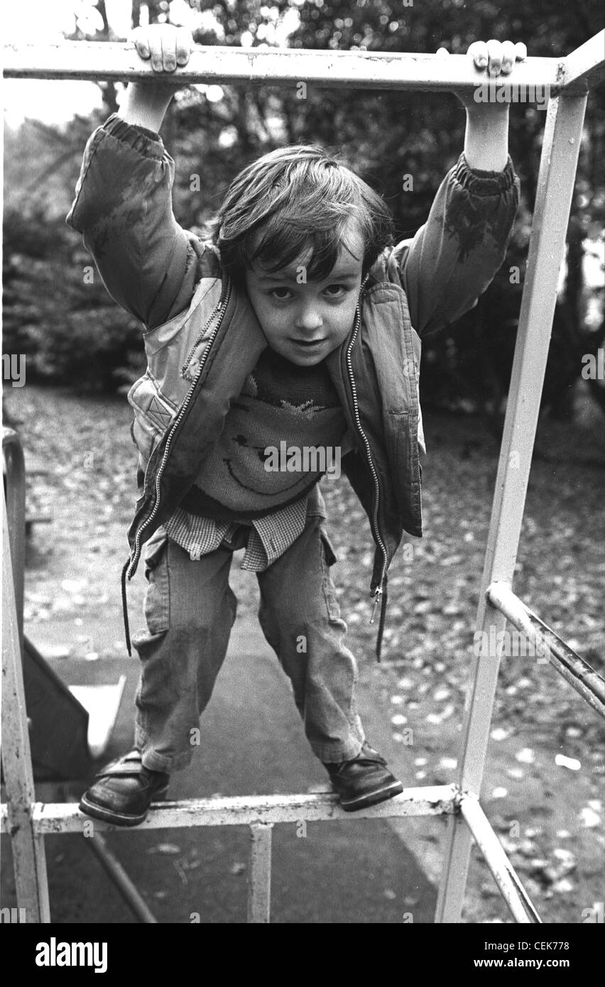 Male child playing on climbing frame in park wearing Whinny The Pooh jumper under hooded jacket arms up looking to camera Stock Photo