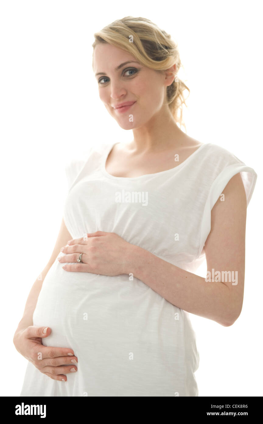 Pregnant female fair hair tied into ponytail, wearing a white dress, standing by the window hands on bump, smiling, looking at Stock Photo