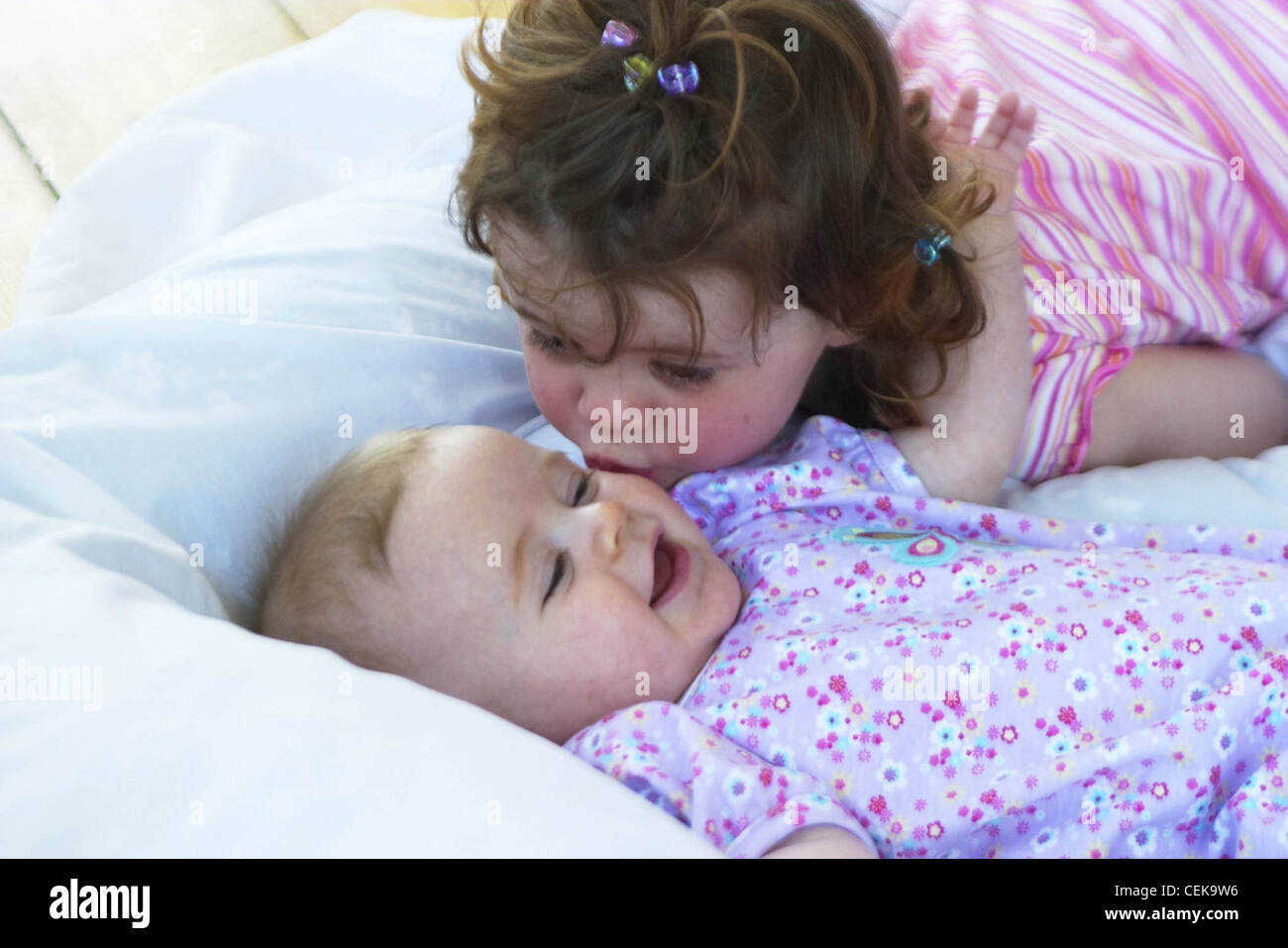 Female child brunette hair tied in topknot wearing pink stripey top leaning over female baby kissing her on cheek, baby is Stock Photo