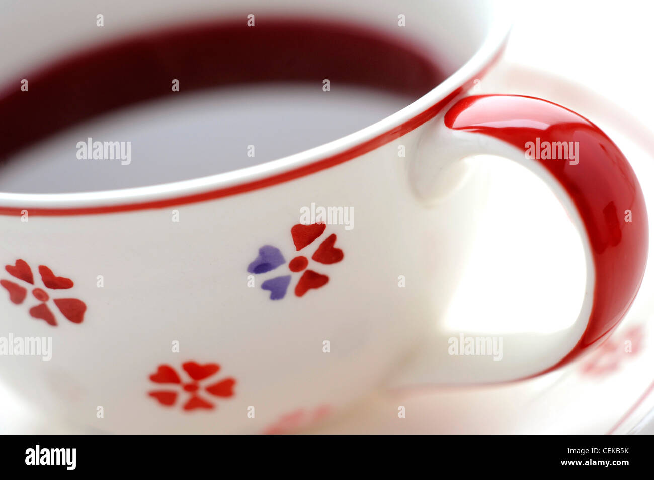 A close up of a white cup and saucer with red flowers on it filled with a red fruit tea Stock Photo