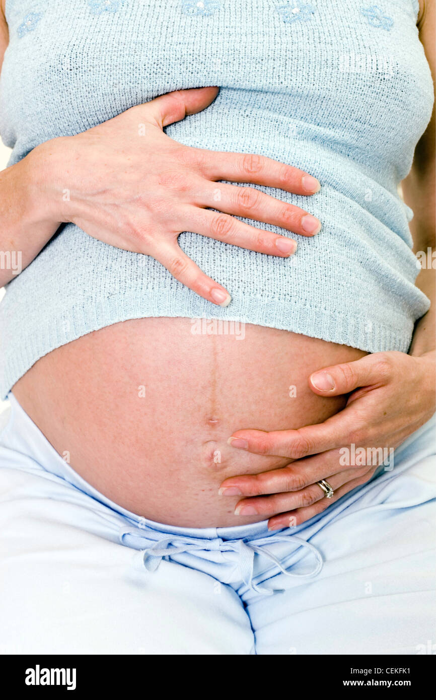 Close up of female wearing pale blue vest and trousers, sitting on bed holding heavily pregnant stomach, straight to camera Stock Photo