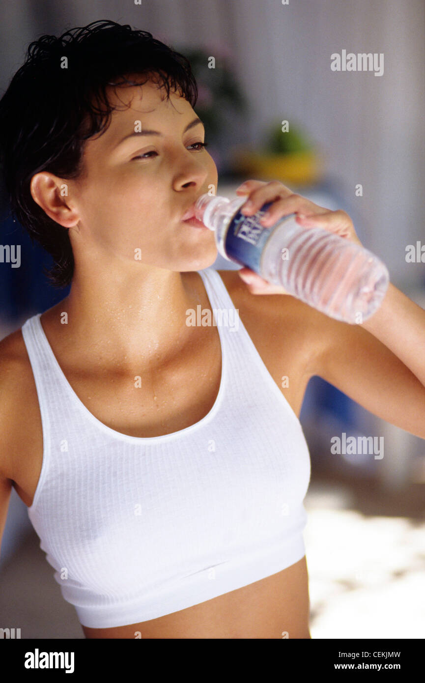 Sweaty brunette female with shirt curly hair wearing a white sports bra and drinking from a water bottle Stock Photo