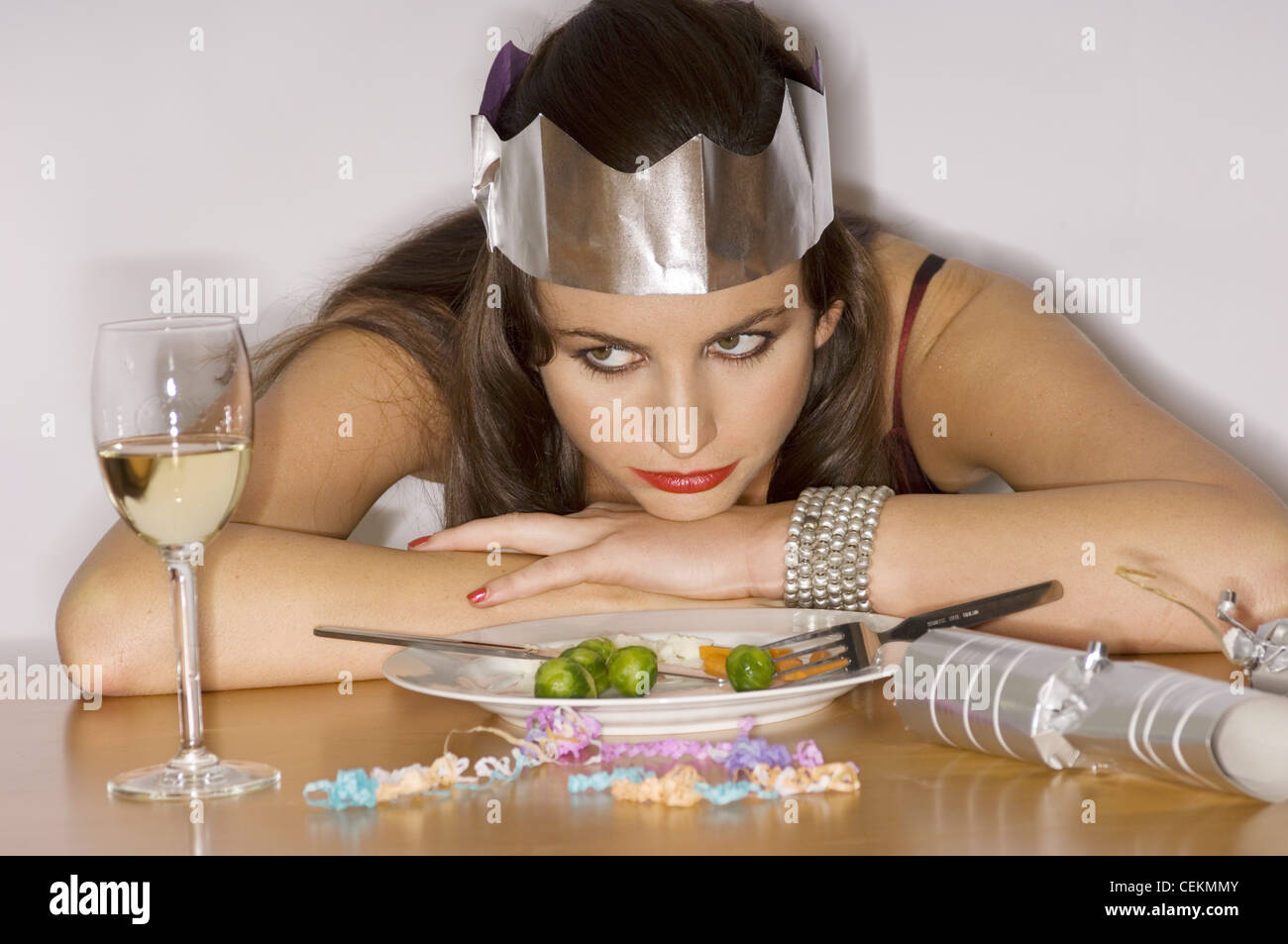 Female brunette hair wearing dark red satin dress, silver paper hat, smokey brown eye make up Sitting at table, elbows on Stock Photo