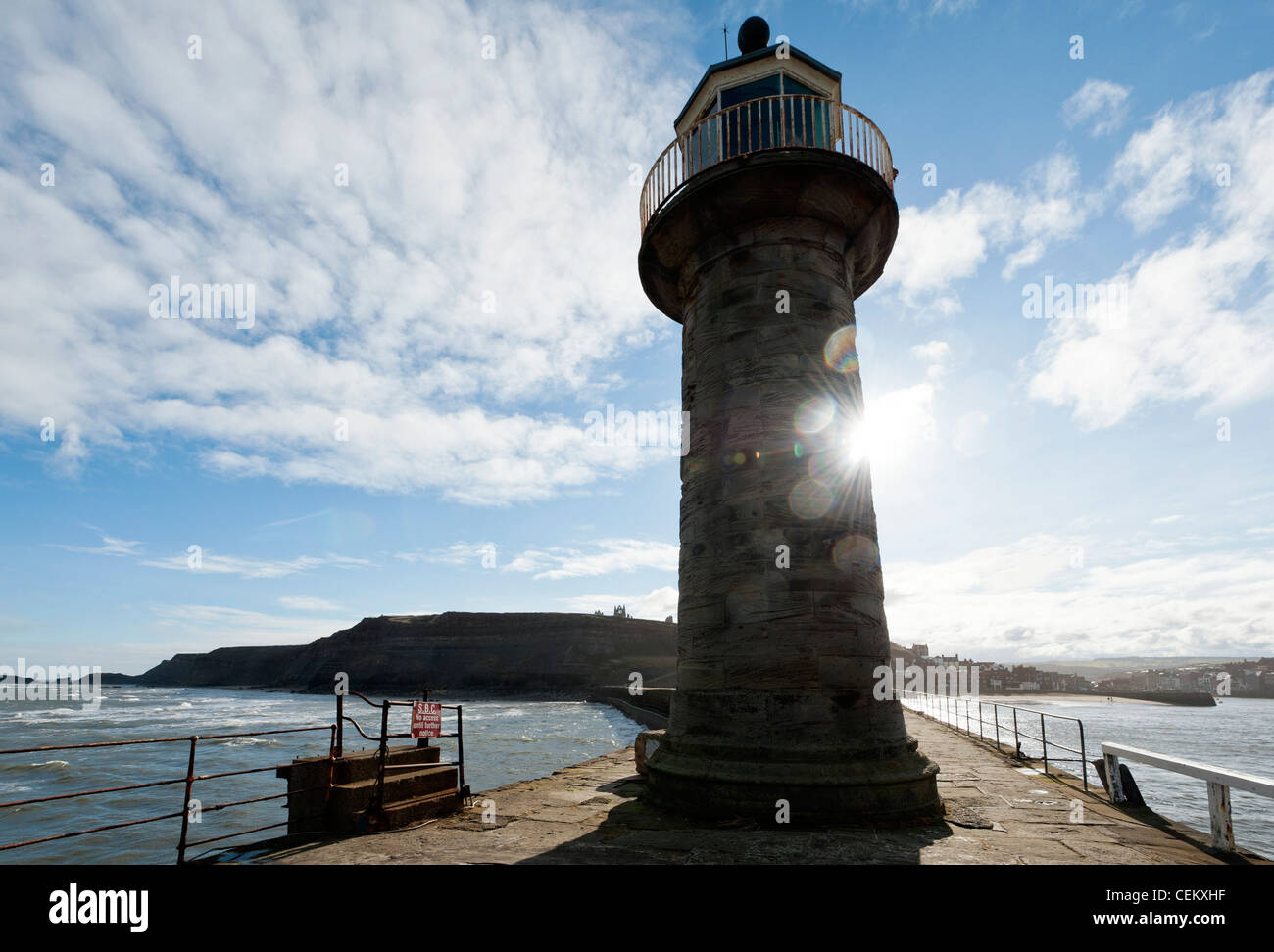 Sun hidden behind Whitby Lighthouse on Pier on a sunny afternoon with a cloudy blue sky Stock Photo