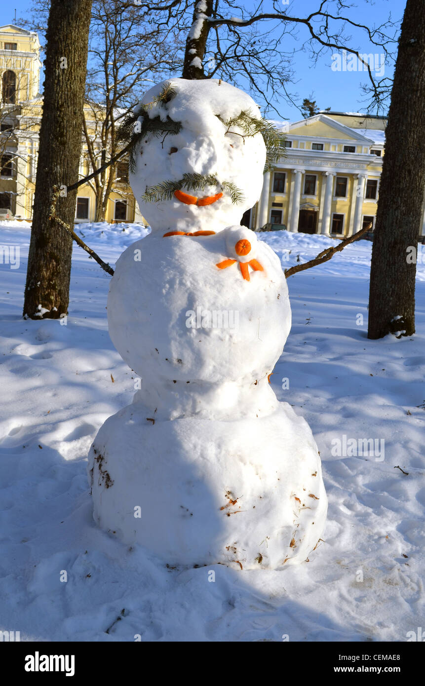 Snowman made of snow decorated with orange bark and spruce branch. Beautiful park view and old palace on background. Stock Photo