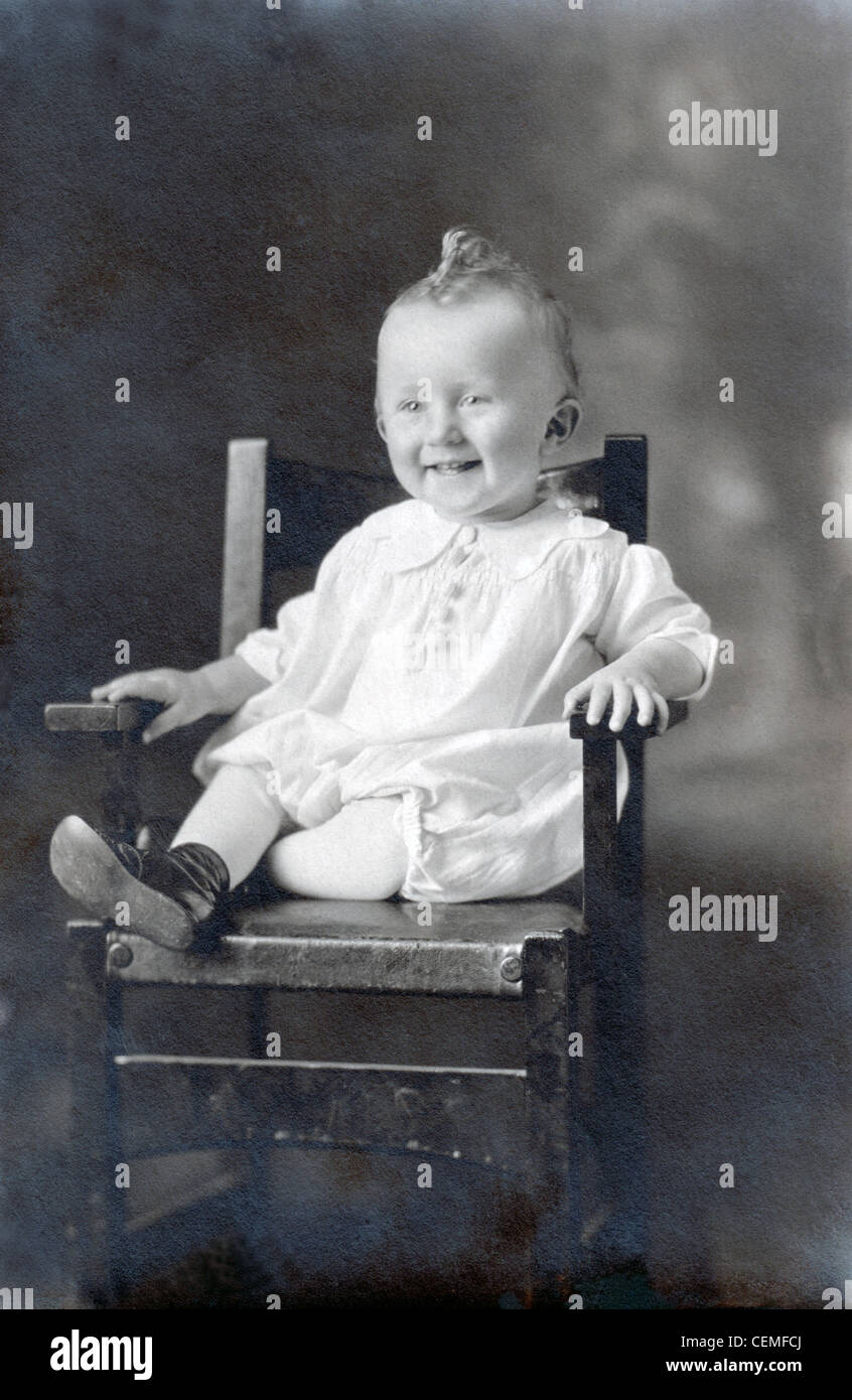 A vintage photograph of a smiling baby with a curl on top of her head, seated in a high chair, circa 1909. Stock Photo