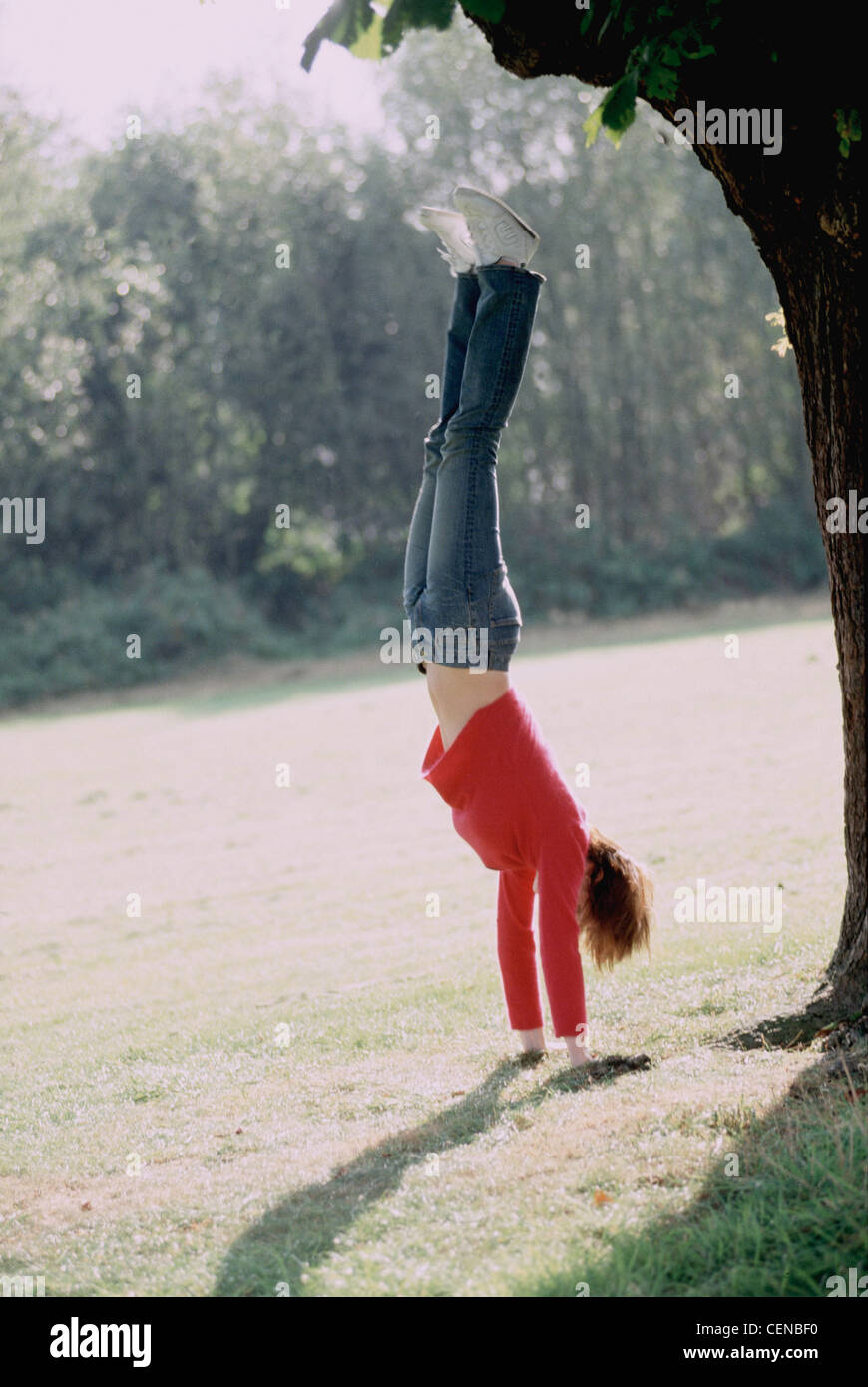A Female with short auburn hair wearing bright pink fluffy jumper blue jeans and trainers, doing a handstand in park Stock Photo