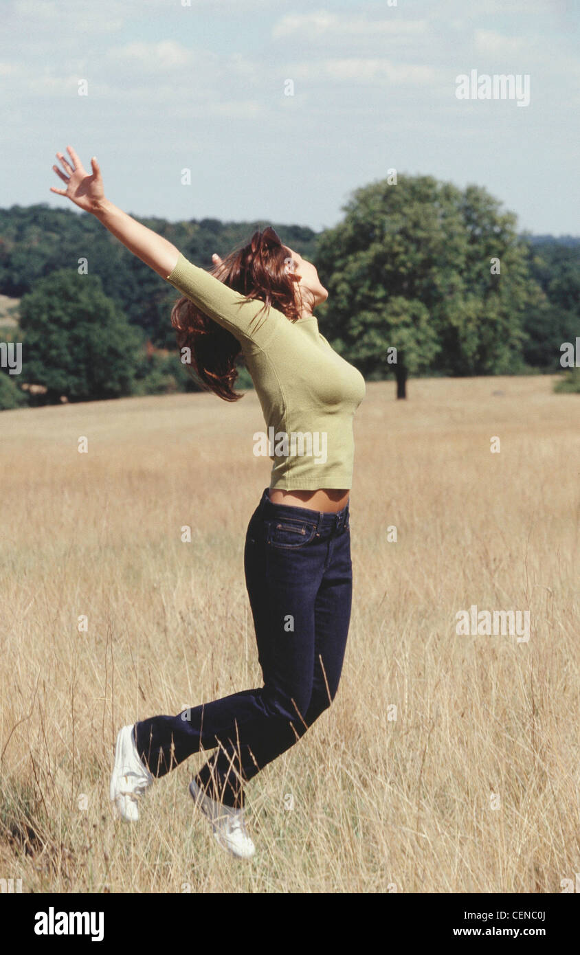 Semi profile of female long brunette hair wearing green top blue jeans and white trainers jumping in air in field arms Stock Photo