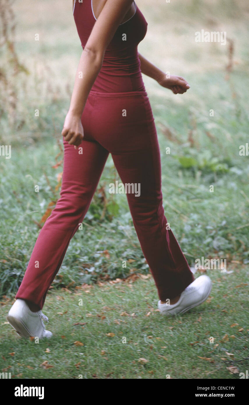 Cropped semi back view of female wearing dark red vest top dark red tracksuit bottoms and white trainers walking in park Stock Photo