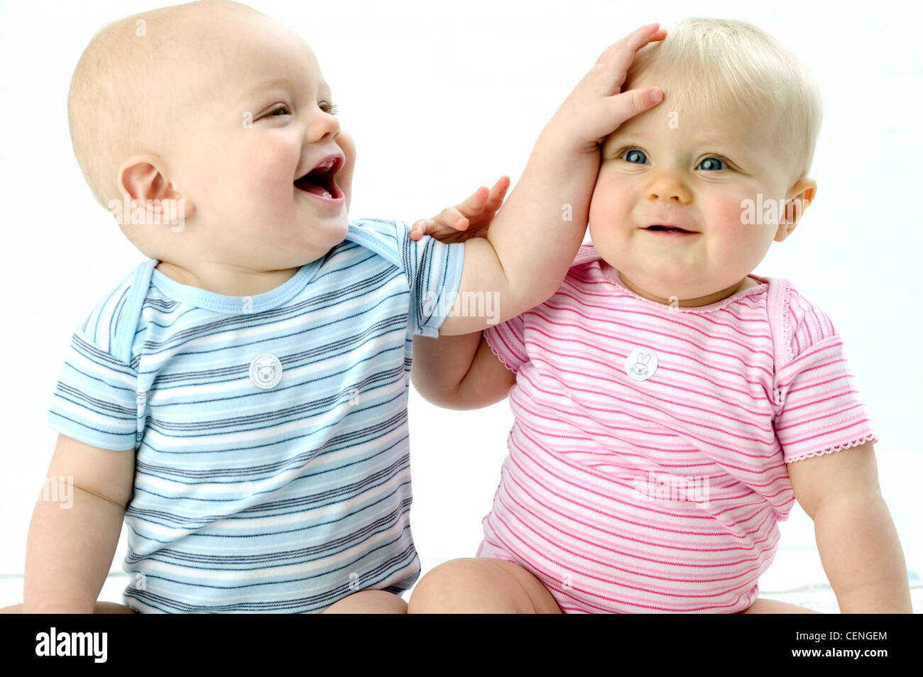 Two babies, male baby on left wearing blue striped babygro looking to side laughing hand on female babys head, baby on right Stock Photo