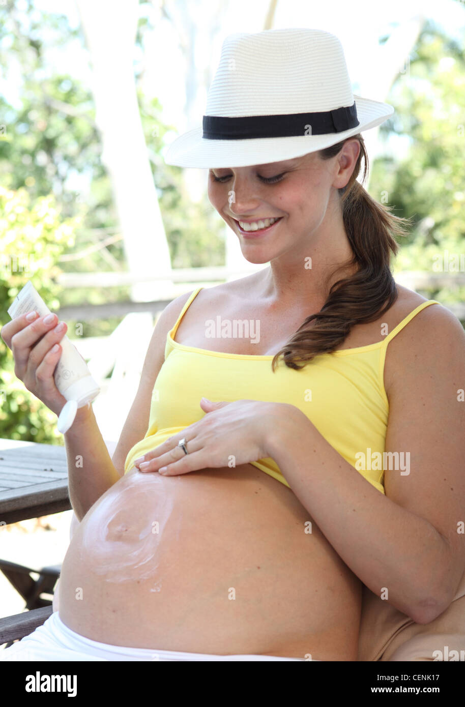 Female long brunette hair, wearing a white hat, white skirt and yellow vest, sitting on armchair, applying cream to her bump, Stock Photo