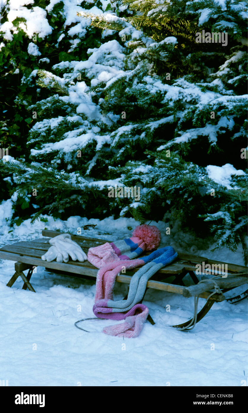 Winter Wonderland Fir trees in the snow with a wood sleigh with woollen gloves, stripey scarf and hat Stock Photo