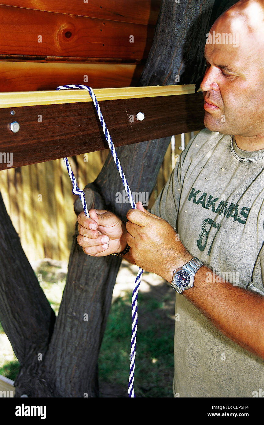 FORBuild a Tree House Step Male tying the rope to the floof the tree house to create a hanging rope ladder Stock Photo
