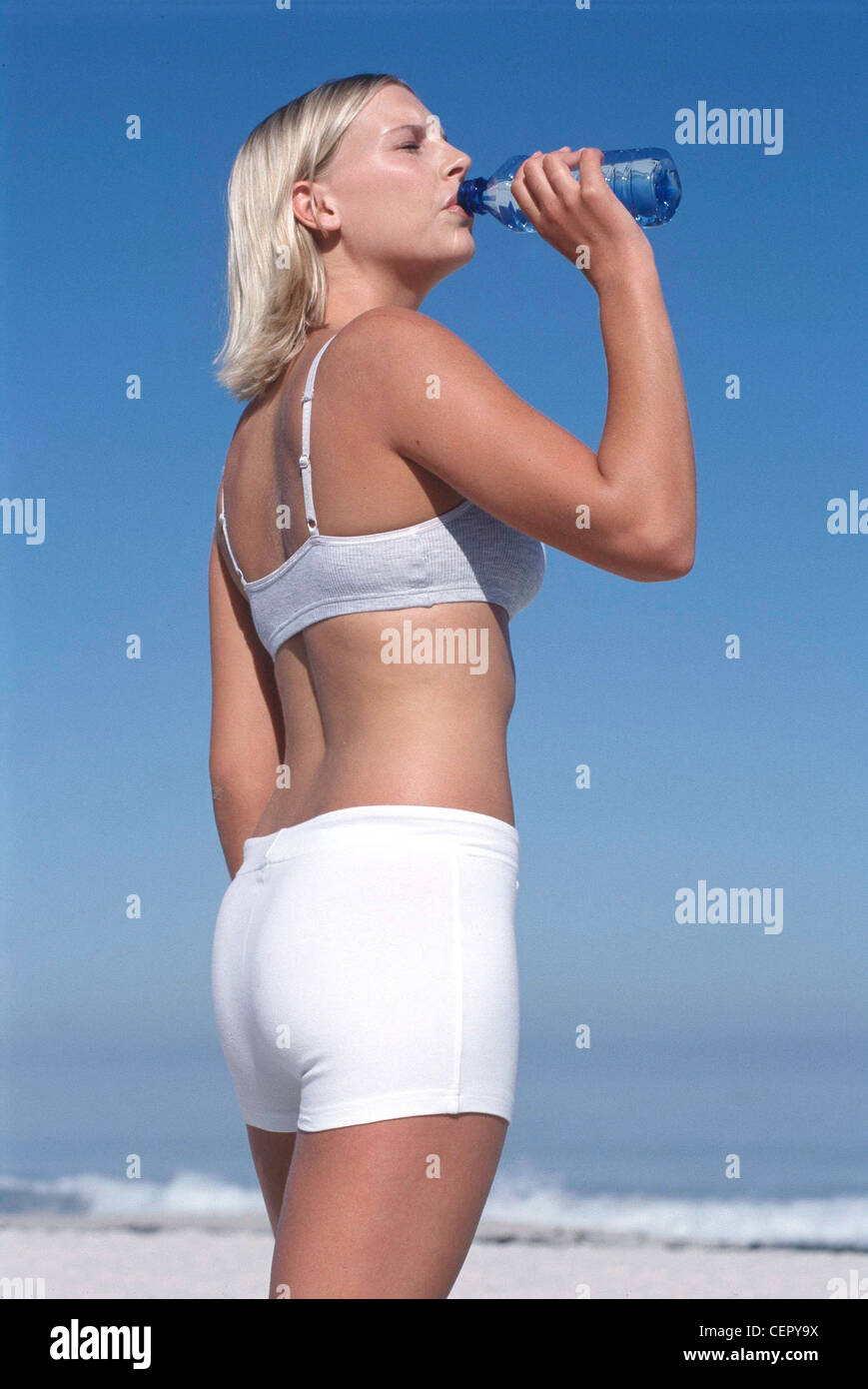 Semi profile of tanned female chin length blonde hair, wearing subtle make up grey top and white shorts, standing on beach by Stock Photo