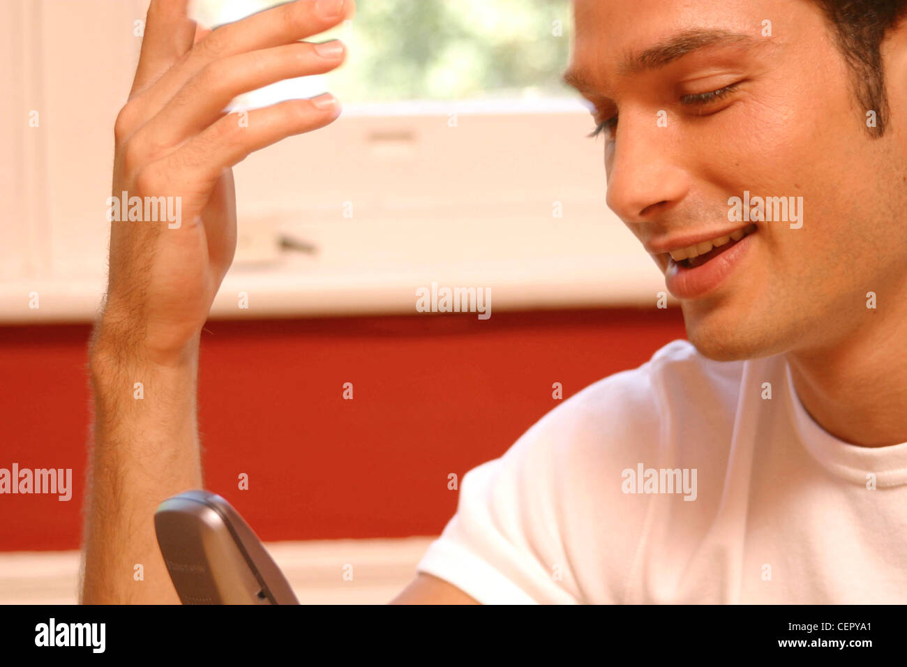 Cropped semi profile of male dark curly hair wearing white t shirt sitting looking at mobile phone one hand raised smiling Stock Photo