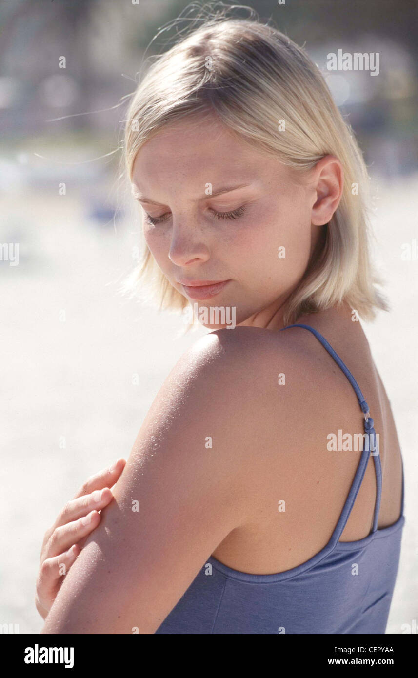 Semi profile of tanned female chin length blonde hair wearing subtle make up and blue vest top, touching arm fingers worried Stock Photo