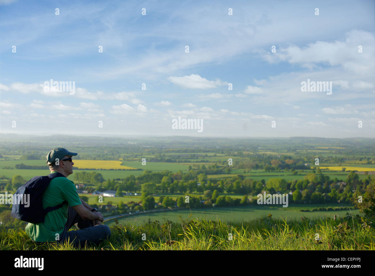A man carrying a rucksack on his back sitting on a hill looking out over the landscape below. Stock Photo
