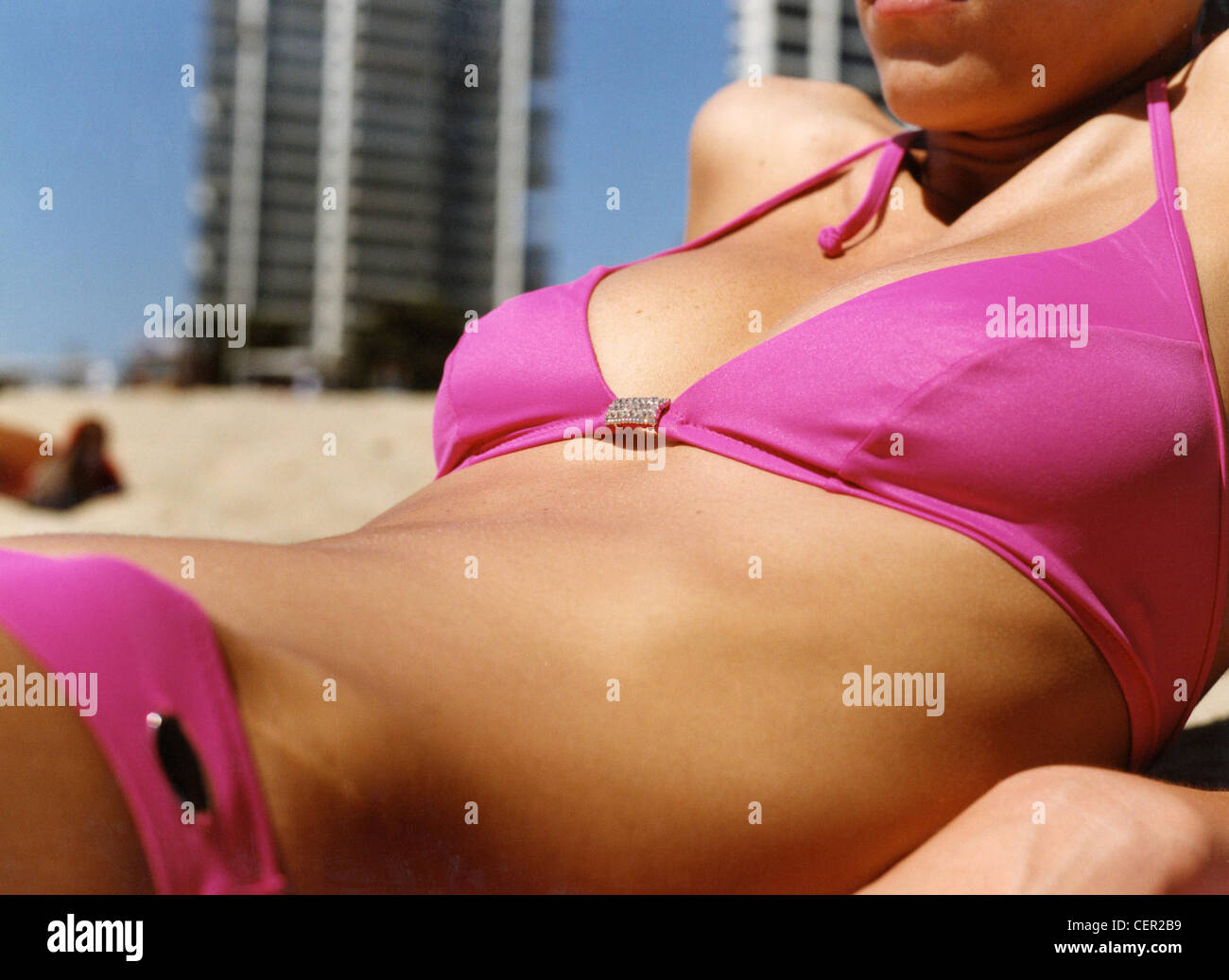 Close up of female chest and stomach, wearing bright pink biknin, lying  back on elbows, beach and buildings in background Stock Photo - Alamy