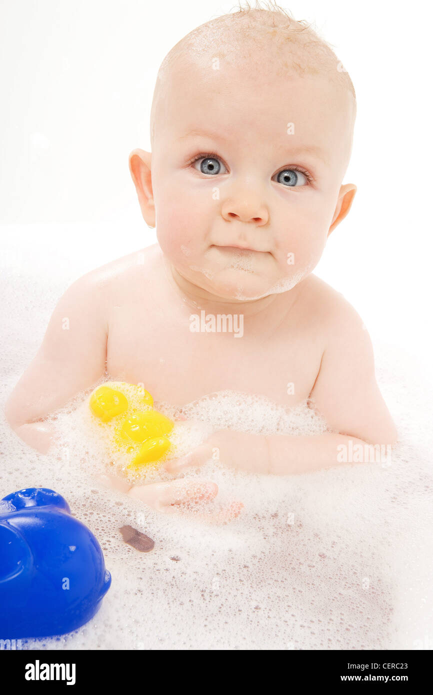 A baby sitting in a bath tub in soapy water with bath toys looking brightly at the camera