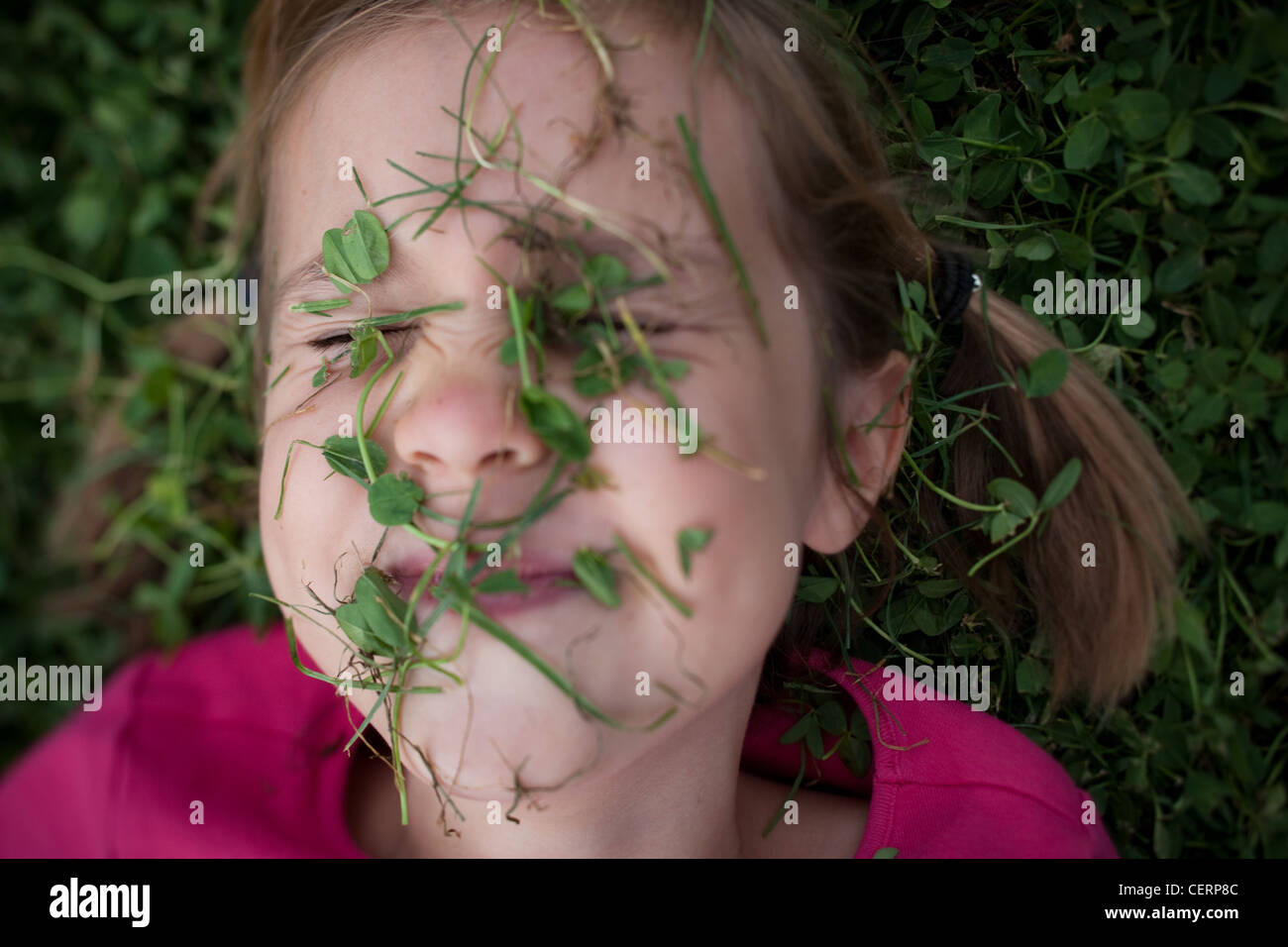 Close up of six year old girl covered in clover leaves and grass, laying on ground, summer. Stock Photo