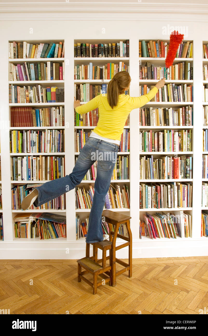 Female standing on step ladder dusting bookcase with red fluffy duster