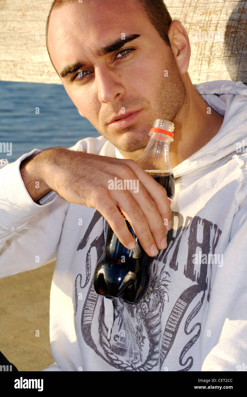 Male short trimmed brunette hair and light stubble, wearing a white hooded top long sleeves, sitting on wooden jetty, drinking Stock Photo