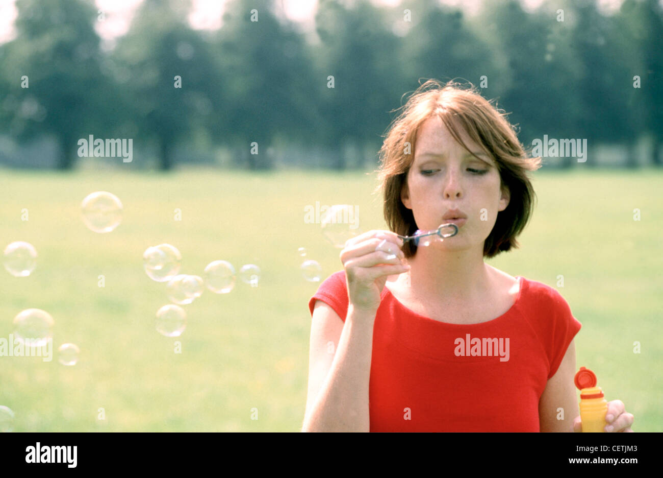 Female with short auburn hair wearing pink lipstick subtle eye make up and red top, blowing bubbles in park Stock Photo