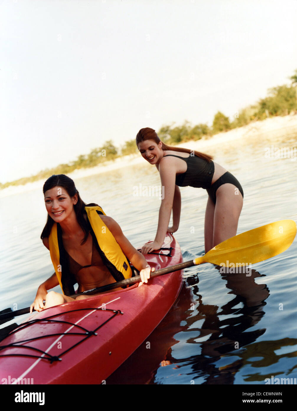 Female long brunette hair, wearing a yellow life vest, sitting in red kayak boat, holding a yellow paddle another female long Stock Photo