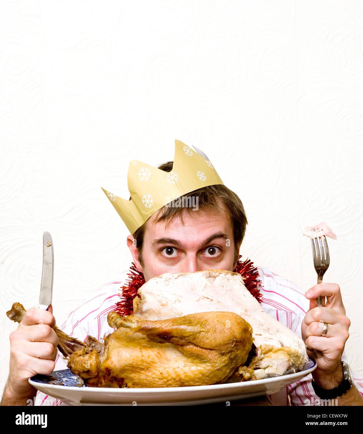 Male wearing paper hat looking out from behind a plate with whole turkey holding knife and fork Stock Photo