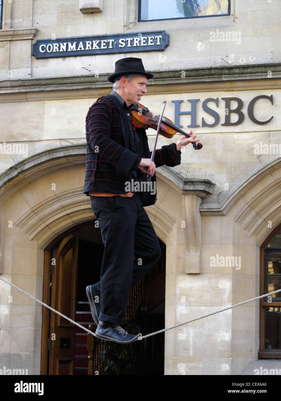 A man playing the fiddle balances on a tightrope outside a bank premises in Oxford High Street. England Stock Photo