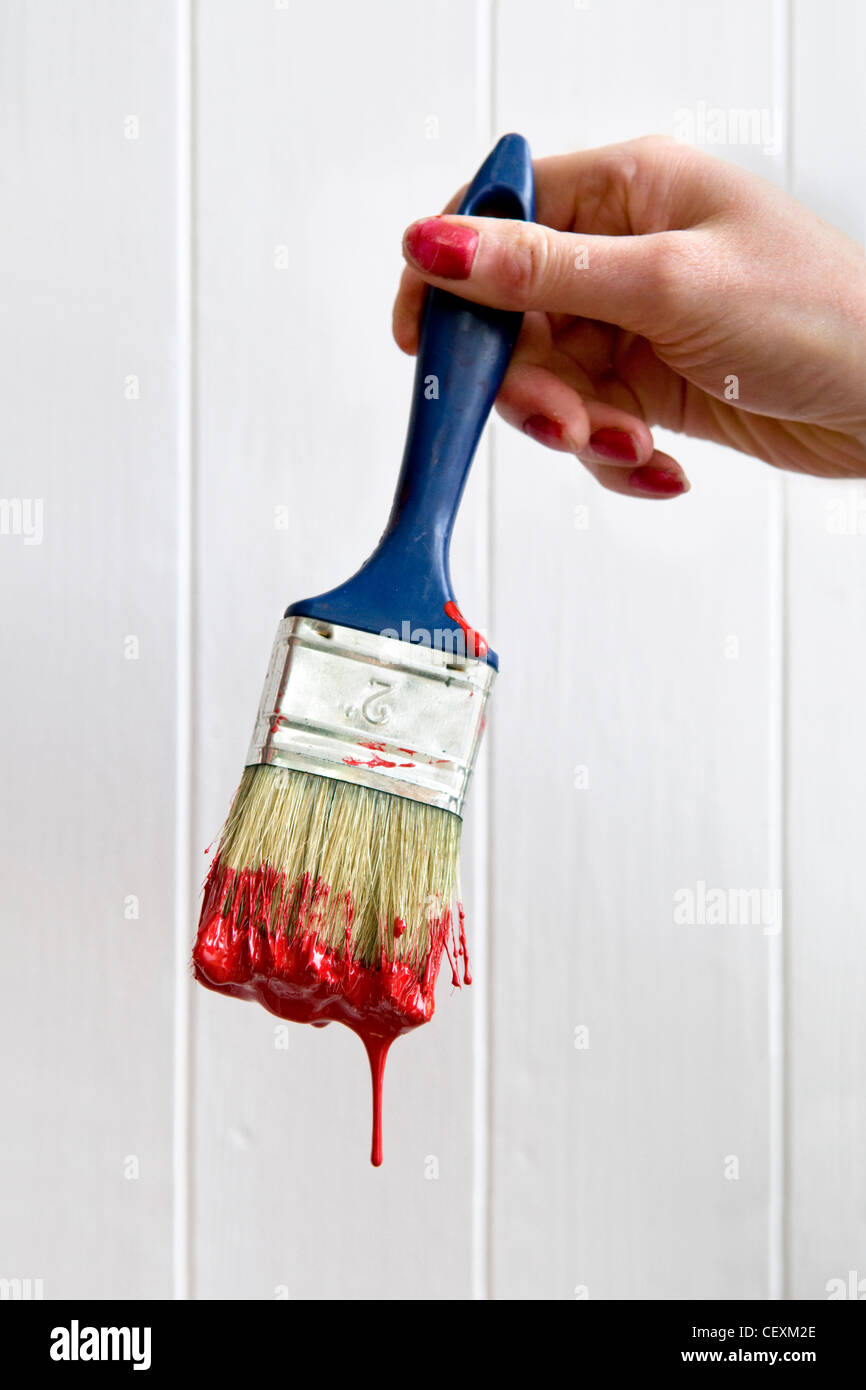 Close up of young Caucasian womans hand holding a dripping paint brush loaded with red paint against a white paneled door Stock Photo