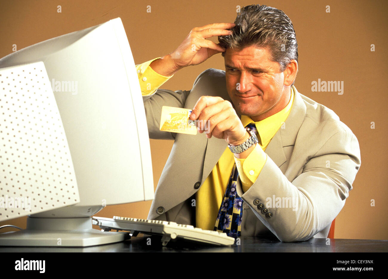 Male wearing pale suit yellow shirt and patterned tie looking at computer monithand to head, holding credit card looking Stock Photo